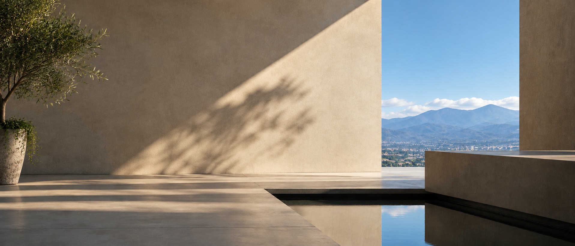 Patio de una casa de muros de estuco al sol, con un olivo en maceta, un espejo de agua y el valle y la montaña al fondo.