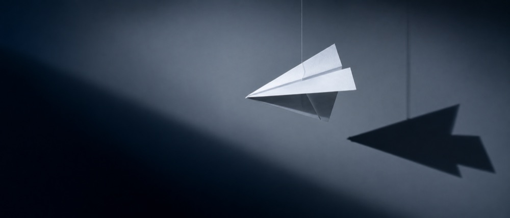 Un avión de papel blanco suspendido de un hilo, con su sombra proyectada en la pared.
